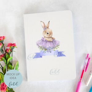 Peut inclure: Un carnet blanc avec une illustration aquarelle d'un lapin brun portant un tutu violet et un ruban bleu. Le texte "on the fold designs" est imprim&eacute; sur le carnet.