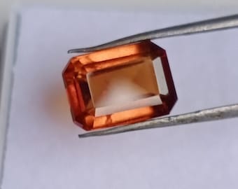 May include: A faceted, rectangular gemstone with a warm orange hue, held by silver tweezers. The gem's cut reveals multiple facets, creating a play of light and shadow. The gem is set against a white background.