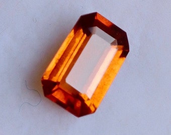 May include: A rectangular, faceted gemstone with a deep orange color. The gemstone is cut in an emerald shape and has a high level of clarity.