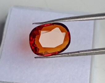 May include: A faceted, oval-shaped gemstone held by tweezers. The gem features a vibrant, reddish-orange hue with a lighter, almost white interior. The gem is set against a white background.