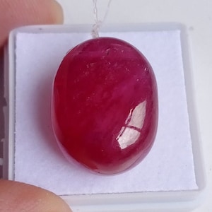 Puede incluir: Una piedra preciosa ovalada de color rojo oscuro con una superficie lisa y pulida. La piedra preciosa se muestra en una caja blanca.