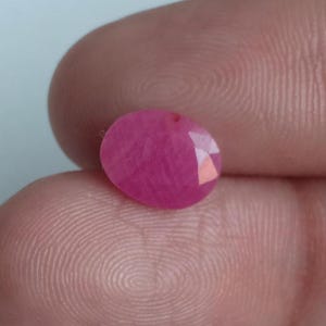 May include: A faceted, oval-shaped ruby gemstone. The gem is a vibrant pinkish-red color, with multiple facets catching the light. The stone is held between two fingers, showcasing its polished surface and internal clarity.