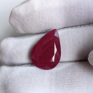以下が含まれることがあります： ファセット加工された、洋梨型のルビーの宝石。ルビーは深く、豊かな赤色で、内部のファセットが見えます。宝石は白い背景に対して保持されています。