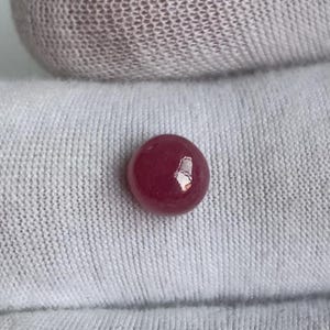 Puede incluir: Primer plano de una gema redonda, pulida, de color rojo intenso, posiblemente un rubí, sobre una tela blanca. La gema tiene un acabado brillante que refleja la luz. La imagen destaca el color y la forma de la gema.