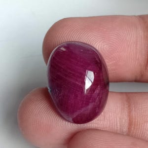 Può includere: Una gemma lucida di forma ovale con una tonalità rosso intenso. La superficie riflette la luce, mostrando striature interne. La gemma è tenuta tra due dita, evidenziando le sue dimensioni e la sua forma. La pietra appare liscia e lucente.