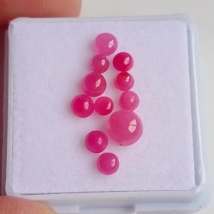 May include: A collection of pink gemstones in a white box. The gemstones are round and smooth, and they are arranged in a random pattern.