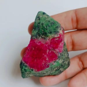 May include: A raw, unpolished mineral specimen featuring a vibrant, deep pinkish-red crystalline centre embedded within a textured, mottled green and black rock matrix. The stone has an irregular, angular shape with a rough, natural surface texture.