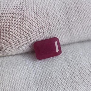 May include: A rectangular, deep red gemstone with a polished surface. The gem has a clear, linear facet running down its centre, reflecting light. The stone is set against a white, textured background.
