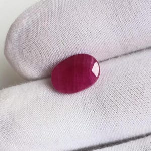 May include: A polished, oval-shaped ruby gemstone. The ruby is a deep, rich red color with visible internal striations. The gemstone is set against a white fabric background, highlighting its color and clarity.