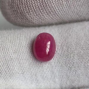 May include: A polished, oval-shaped ruby gemstone. The gem is a deep, vibrant red color with a glossy surface. The ruby is set against a white, textured background.