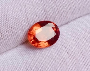 May include: An oval-cut, orange-red gemstone with a faceted surface. The gem is set against a white fabric background, highlighting its color and clarity. The gemstone's facets reflect light, creating a sparkling effect.