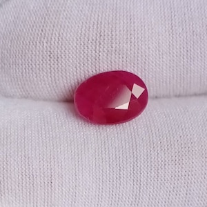 Può includere: Una gemma ovale di colore rosso vibrante con taglio sfaccettato. La gemma è posizionata su uno sfondo bianco.