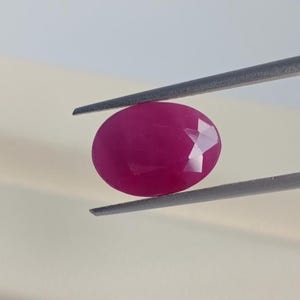 Op de afbeelding: Een gefacetteerde, ovaalvormige robijn edelsteen, vastgehouden door een pincet. De robijn heeft een diepe, verzadigde rode kleur, met meerdere facetten die het licht vangen. De pincet is zilverkleurig, tegen een neutrale achtergrond.