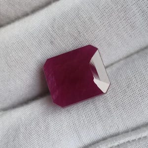 May include: A loose, rectangular-cut gemstone with a deep, saturated magenta-red color. The stone features faceted surfaces that create distinct light reflections and shadows across its top, showcasing a clear, polished, and translucent appearance.