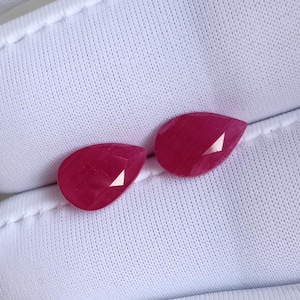 May include: Two faceted, teardrop-shaped, ruby-red gemstones. The gems have a polished surface with triangular facets that reflect light. The stones are displayed on a white, textured fabric.