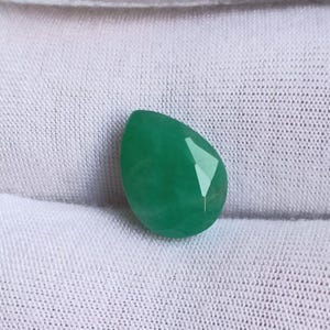 May include: A faceted, teardrop-shaped gemstone in a vibrant green hue. The gem is set against a white fabric background, highlighting its polished surfaces and color. The gem's cut creates multiple reflective facets.