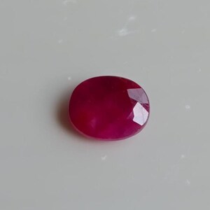 Puede incluir: Una gema de rubí ovalada y facetada. La gema es de color rojo intenso y saturado, con una superficie pulida y facetas visibles. La piedra está sobre un fondo blanco.