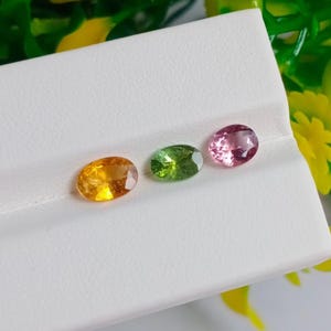 May include: Three oval gemstones in shades of yellow, green, and pink are displayed on a white surface. The gemstones are polished and reflect light, showcasing their clarity and color. The arrangement highlights the variety of colors available.