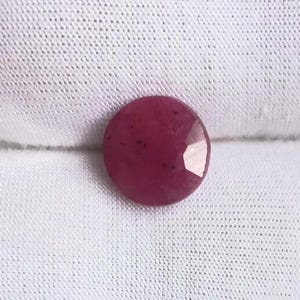 May include: A faceted, round, deep red gemstone, possibly a ruby, rests on a white fabric. The gem's surface reflects light, showcasing its cut. The stone's color is a rich, saturated red, with subtle variations in tone.