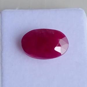 Peut inclure: Une pierre précieuse de rubis ovale et facettée. Le rubis est d'un rouge profond et riche, avec une surface polie et un bord facetté qui capte la lumière. La pierre est posée sur un fond blanc.