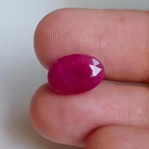 以下が含まれることがあります： ファセット加工された、楕円形のルビーの宝石。ルビーは、研磨された表面を持つ、深く鮮やかな赤色をしています。宝石は2本の指の間に挟まれ、そのサイズと輝きを示しています。石のカットは、光を反射する複数のファセットを作成します。