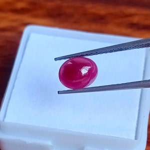 May include: A vibrant red oval-shaped gemstone held by tweezers. The gemstone is smooth and polished, reflecting light.