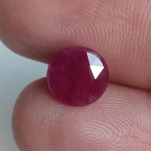 Pode incluir: Uma pedra preciosa redonda e facetada com uma cor vermelho-magenta profunda e saturada. A pedra apresenta uma superfície polida com múltiplas facetas triangulares que refletem a luz, criando um brilho intenso num lado da pedra.
