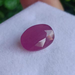 Puede incluir: Una gema de rubí facetada de forma ovalada. La gema es de color magenta intenso con una superficie pulida y algunas facetas que reflejan la luz. El rubí está sobre un fondo blanco.