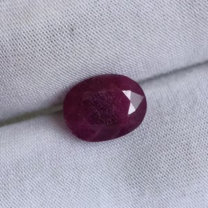 May include: A faceted, oval-shaped ruby gemstone. The gem is a deep, rich red color with visible internal inclusions. The stone is set against a white fabric background, highlighting its color and cut.