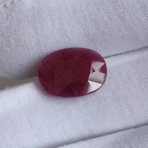 Pode incluir: Uma pedra preciosa de rubi oval e facetada. A gema é de uma cor vermelha profunda e rica com múltiplas facetas que capturam a luz. O rubi está sobre um fundo de tecido branco, destacando sua cor e clareza.