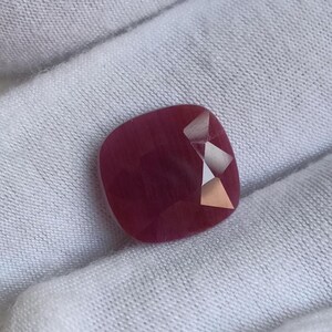 May include: A cushion-cut gemstone with a deep, saturated magenta-red hue. The stone features a smooth, polished surface with a distinct faceted section on the right side that reflects light, creating sharp, bright geometric highlights.