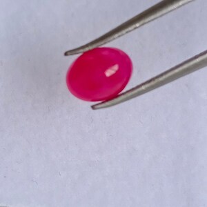 Puede incluir: Una piedra preciosa ovalada de color rosa brillante sostenida por unas pinzas.