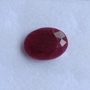 May include: A faceted, oval-shaped ruby gemstone. The gem is a deep, rich red color with multiple facets that catch the light. The gemstone is set against a plain white background.