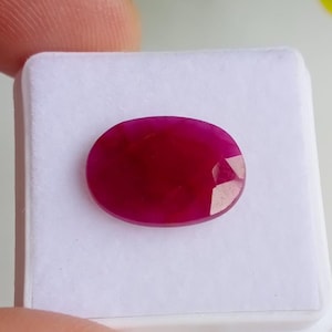 Puede incluir: Una gema de rubí facetada de forma ovalada. El rubí es de un color rojo intenso y saturado, con una superficie pulida. La gema está sobre un fondo blanco.