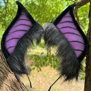 May include: A pair of black and purple bat wing ears on a headband. The wings are black with purple ribbed detailing. The ears are made of faux fur and are attached to a thin, black headband.