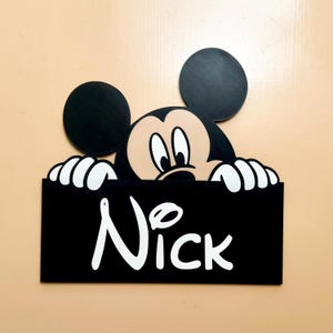 May include: Black and white wooden sign with a Mickey Mouse character peeking over the top. The sign says "Nick" in white letters.