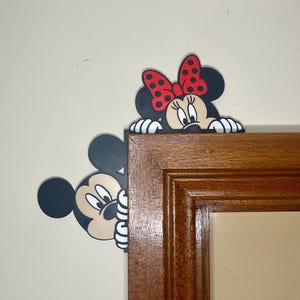 May include: Mickey Mouse and Minnie Mouse peeking around the corner of a wooden picture frame. Mickey is on the left and Minnie is on the right. Mickey is black with large ears and Minnie has a red bow with white polka dots.