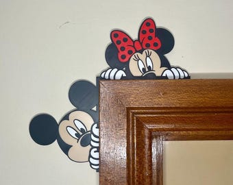 May include: Mickey Mouse and Minnie Mouse peeking around the corner of a wooden picture frame. Mickey is on the left and Minnie is on the right. Mickey is black with large ears and Minnie has a red bow with white polka dots.
