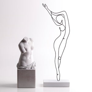 May include: A white plaster bust of a woman's torso on a white pedestal next to a black wire sculpture of a woman on a white pedestal.