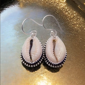 Puede incluir: Un par de pendientes en forma de lágrima con un diseño de concha natural. Los pendientes tienen un borde de metal plateado con pequeños detalles de cuentas. Las conchas son de color blanco cremoso.