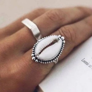 Puede incluir: Un anillo de plata con un diseño de concha blanca. El anillo está en un dedo y el fondo es un libro.