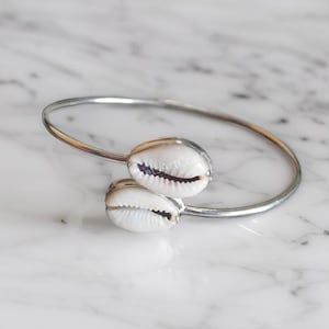 May include: Silver bangle bracelet with two white cowrie shells attached. The bracelet is open and the shells are facing the viewer.