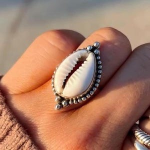 Puede incluir: Un anillo de plata con un diseño de concha blanca. El anillo está adornado con pequeñas cuentas de plata alrededor del borde de la concha.