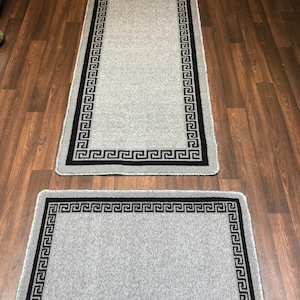 May include: Two grey rugs with black Greek key borders. The larger rug is a runner and the smaller rug is a square.