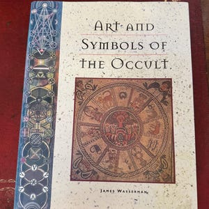 May include: A hardcover book titled "Art and Symbols of the Occult" by James Wasserman. The cover features a circular illustration with various symbols and figures in shades of brown and red. The spine has a geometric design in blue and green.
