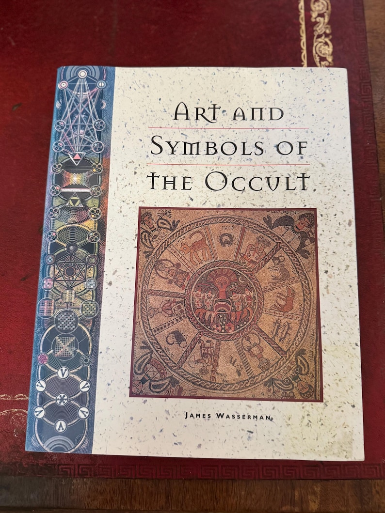 May include: A hardcover book titled "Art and Symbols of the Occult" by James Wasserman. The cover features a circular illustration with various symbols and figures in shades of brown and red. The spine has a geometric design in blue and green.
