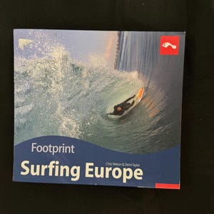 May include: A book cover with a surfing theme. The cover features a surfer riding a wave inside a barrel. The book title is "Footprint Surfing Europe" in white and blue text. A red square with a footprint logo is in the upper right corner.