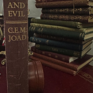 May include: A collection of vintage books in various colours and conditions. The prominent book's spine displays "GOD AND EVIL" and "C.E.M. JOAD" in gold lettering. A small, brown leather case is also visible.