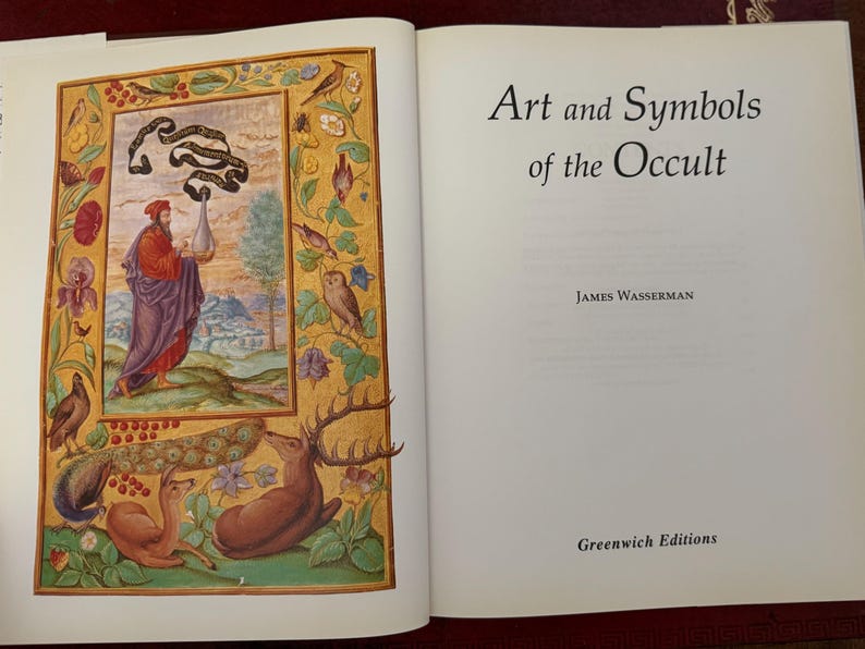May include: An open book with the title "Art and Symbols of the Occult" by James Wasserman. The left page features a colorful illustration with a figure in red and purple robes, surrounded by birds, deer, and floral elements.