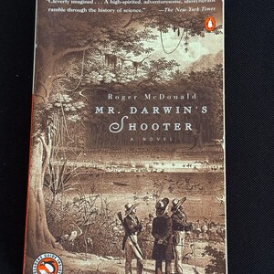 May include: A paperback book titled "Mr. Darwin's Shooter: A Novel" by Roger McDonald. The cover features a sepia-toned illustration of figures in period clothing near a body of water. The Penguin Readers Guide logo is visible.